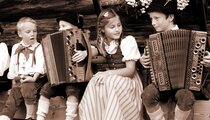 Kinderen in traditionele alpenkleding zitten voor een houten hut, terwijl twee van hen op een trekzak spelen en regionale muziektradities uitbeelden. | © salzburgerland.at