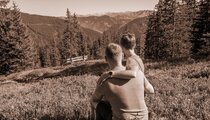 Twee personen zitten samen op een bergweide en genieten van het weidse uitzicht over bossen en het omliggende berglandschap. | © viehhofen.at, Der Fotoigel
