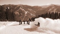 Skiër rijdt door verse poedersneeuw op een besneeuwde skipiste, terwijl andere wintersporters wachten tegen een alpien bergdecor. | © viehhofen.at, Der Fotoigel