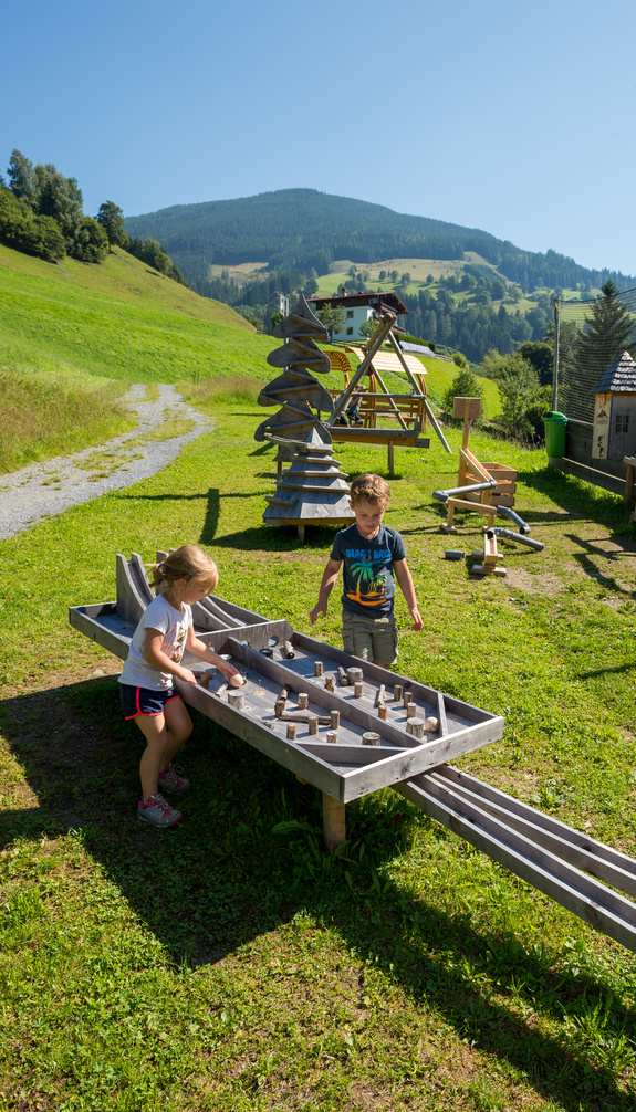Two children play at a large marble run in Viehhofen, surrounded by meadows and other play elements against a green mountain backdrop. | © viehhofen.at, Der Fotoigel