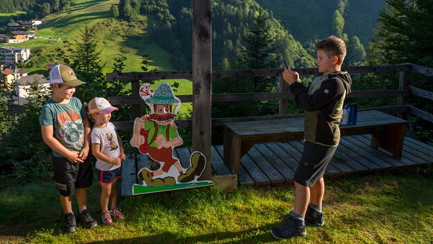 Ein Kind fotografiert zwei Kinder gemeinsam mit Wander-Nik an einer Erlebnisstation oberhalb von Viehhofen, mit Blick auf den Ort und die grüne Berglandschaft. | © viehhofen.at, Der Fotoigel