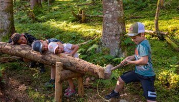 Four children explore a wooden activity station on the children’s hiking trail in Viehhofen, with one child striking a log with a wooden mallet while the others watch. | © viehhofen.at, Der Fotoigel