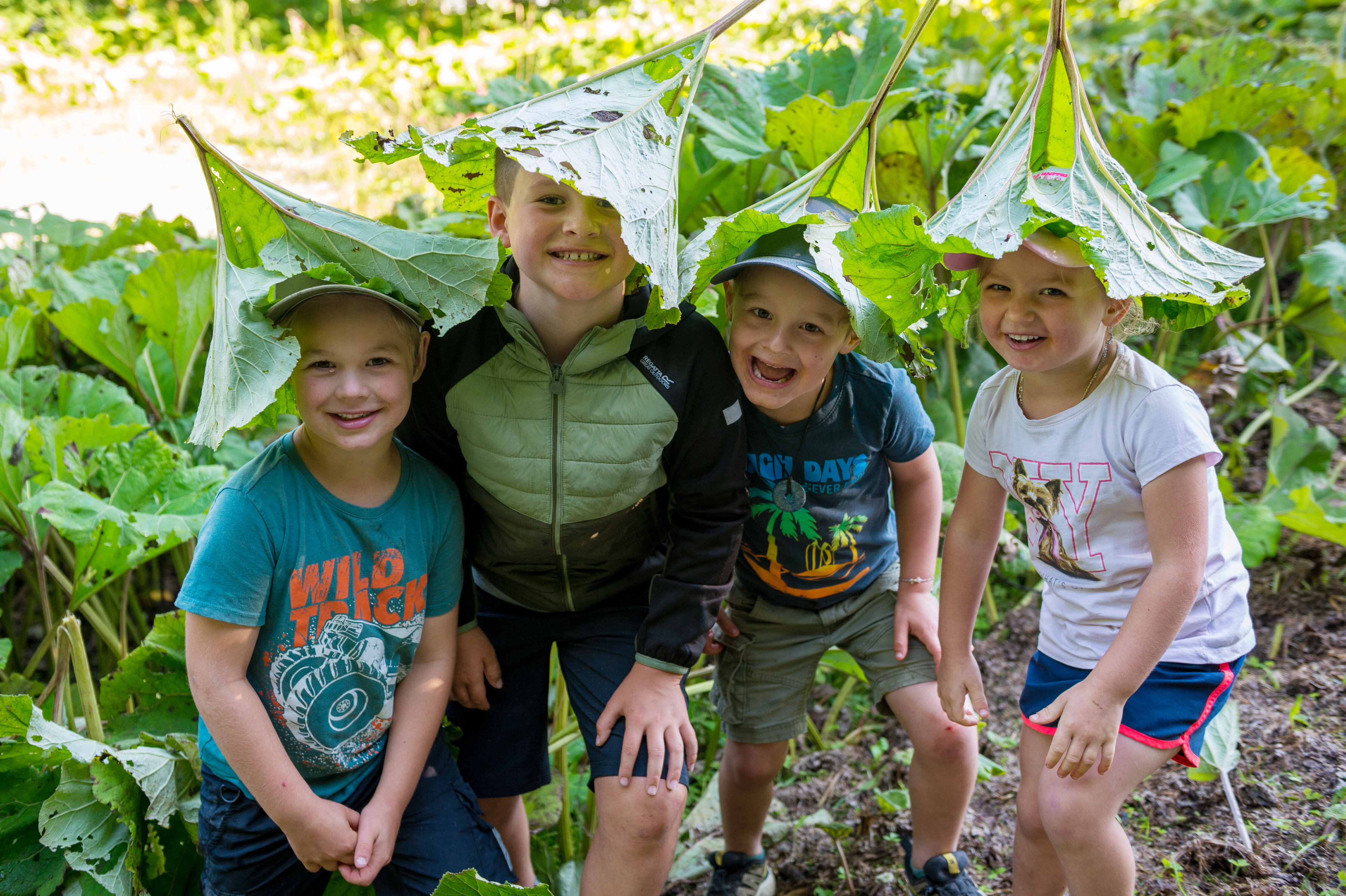 Four children on the children’s hiking trail in Viehhofen laughing beneath large green leaves they are playfully wearing like hats. | © viehhofen.at, Der Fotoigel