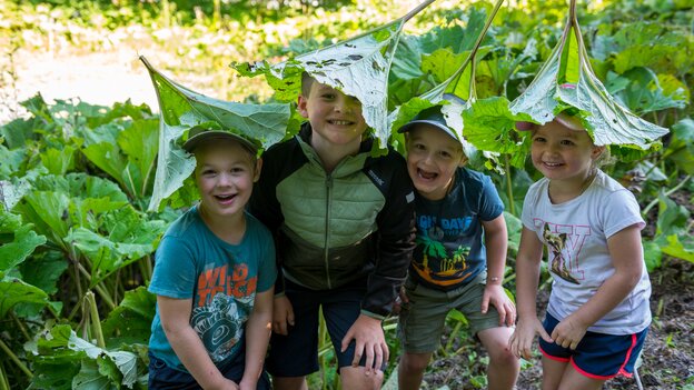 Vier Kinder am Kinderwanderweg in Viehhofen lachen unter großen grünen Blättern, die sie spielerisch wie Hüte über dem Kopf tragen. | © viehhofen.at, Der Fotoigel