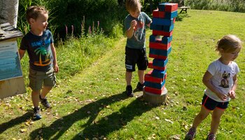 Three children playing at the Jenga activity station in Viehhofen with large red and blue wooden blocks on a grassy meadow. | © viehhofen.at, Der Fotoigel