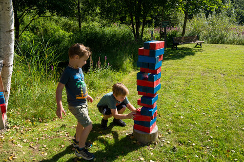 Two children play at a Jenga activity station in Viehhofen, carefully removing wooden blocks from a tall red and blue tower on a grassy meadow. | © viehhofen.at, Der Fotoigel