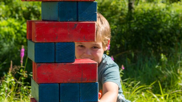 Kind spielt an einer Jenga-Spielstation in Viehhofen und zieht vorsichtig einen Holzklotz aus einem hohen rot-blauen Turm. | © viehhofen.at, Der Fotoigel
