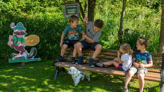 Vier Kinder machen mit einem kleinen Hund auf einer Holzbank in Viehhofen Pause, neben einer Wander-Nik-Figur und umgeben von grüner Natur. | © viehhofen.at, Der Fotoigel
