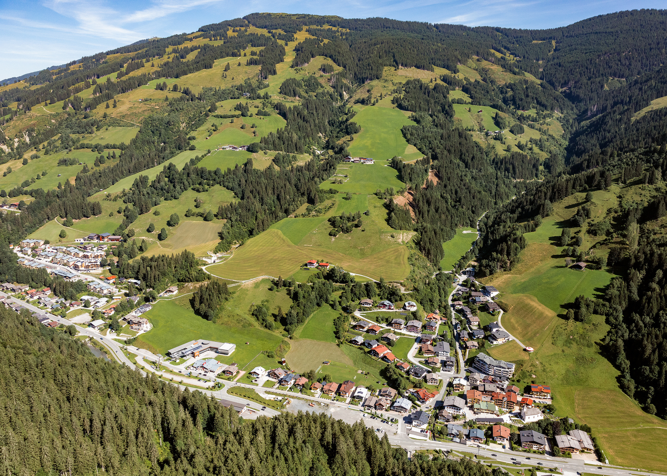 Luftaufnahme von Viehhofen im Sommer mit dem Ortsgebiet im Tal, grünen Wiesen, bewaldeten Berghängen und der umliegenden Berglandschaft im Glemmtal. | © viehhofen.at, Amido