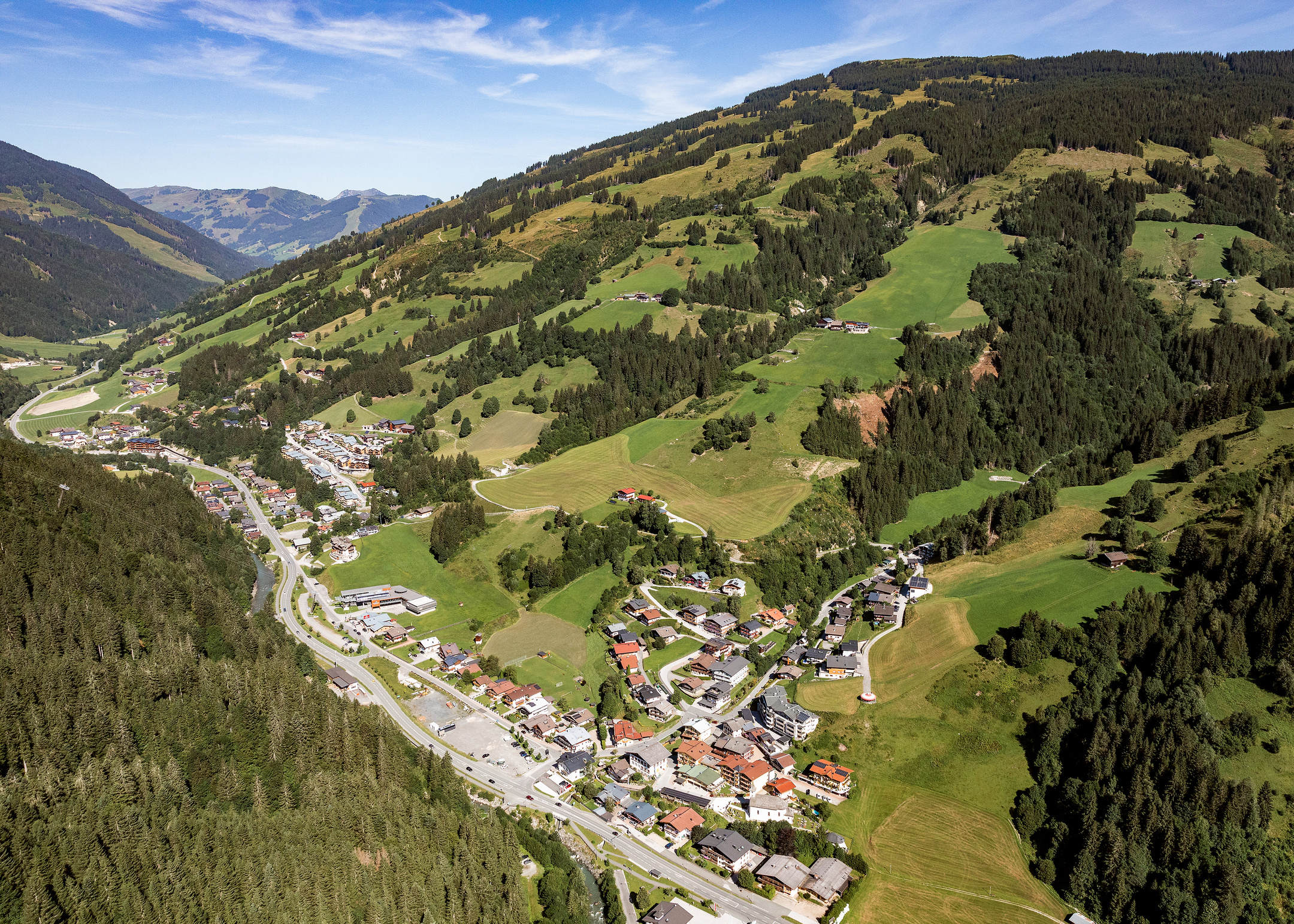 Luftaufnahme von Viehhofen im Sommer mit dem Ortsgebiet im Tal, grünen Wiesen, bewaldeten Berghängen und der umliegenden Berglandschaft im Glemmtal. | © viehhofen.at, Amido