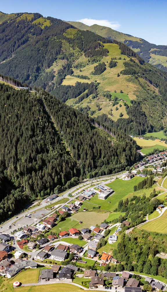 Luftaufnahme von Viehhofen im sommerlichen Glemmtal mit dem Ort im Talboden, grünen Wiesen, dicht bewaldeten Berghängen und der umliegenden Berglandschaft. | © viehhofen.at, Amido