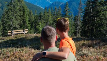 Paar sitzt eng umschlungen auf einer Bergwiese in Viehhofen und blickt über Fichtenwald auf die umliegende Bergwelt. | © viehhofen.at, Der Fotoigel
