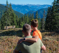 Couple sitting close together on a mountain meadow in Viehhofen, looking across spruce forest towards the surrounding mountains. | © viehhofen.at, Der Fotoigel