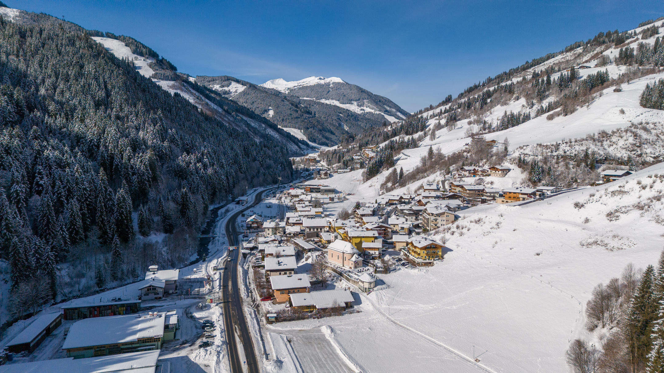 Luftaufnahme von Viehhofen im Winter mit verschneitem Ortsgebiet, weißen Wiesen, Talstraße und bewaldeten Berghängen im Glemmtal. | © viehhofen.at, Der Fotoigel