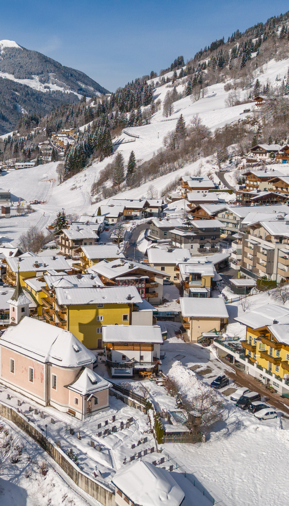 Luftaufnahme von Viehhofen im Winter mit verschneitem Ortsgebiet, weißen Wiesen, Talstraße und bewaldeten Berghängen im Glemmtal. | © viehhofen.at, Der Fotoigel