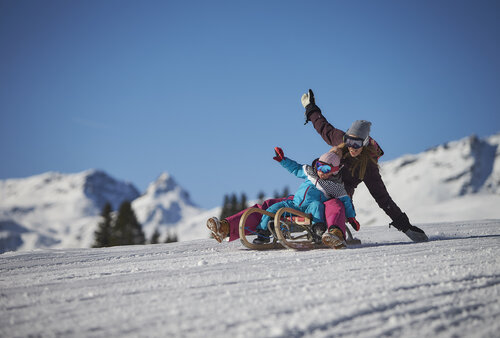 Erwachsene Person und Kind beim Rodeln auf einer verschneiten Piste in Saalbach, mit winterlicher Berglandschaft im Hintergrund. | © viehhofen.at, saalbach.com