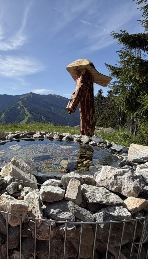 Glücksbrunnen oberhalb von Viehhofen mit Holz-Wasserspeier, Wasserbecken und bunten Schmucksteinen vor der umliegenden Bergkulisse. | © viehhofen.at