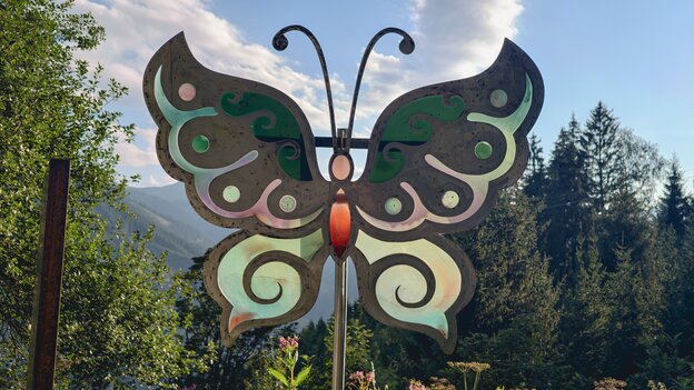 Buntglas-Schmetterling am Glücksweg in Viehhofen, von der Sonne hinterleuchtet und umgeben von Wildblumen, Bäumen und Berglandschaft. | © viehhofen.at