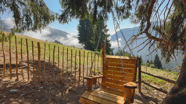 Hölzerner Hängesessel unter einem großen Baum am Glücksweg in Viehhofen, mit Blick auf Wiesen und die umliegende Berglandschaft.