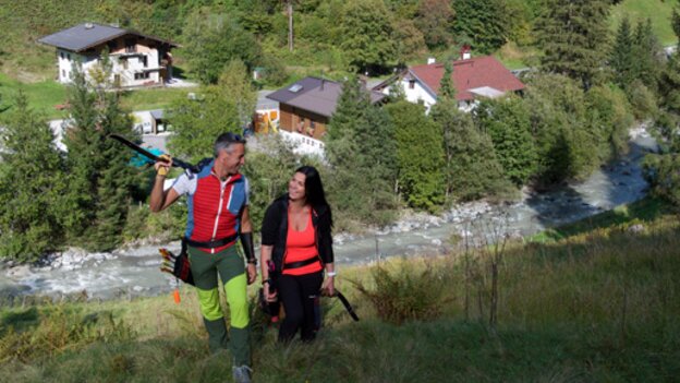 Two people walking along the archery course in Viehhofen with bows and equipment, with green hillsides and houses in the valley behind them. | © Der Fotoigel