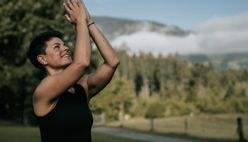 Eine Frau macht Yogaübungen in der Natur. Im Hintergrund sieht man einen Wald und die Bergwelt | © Anna Überegger